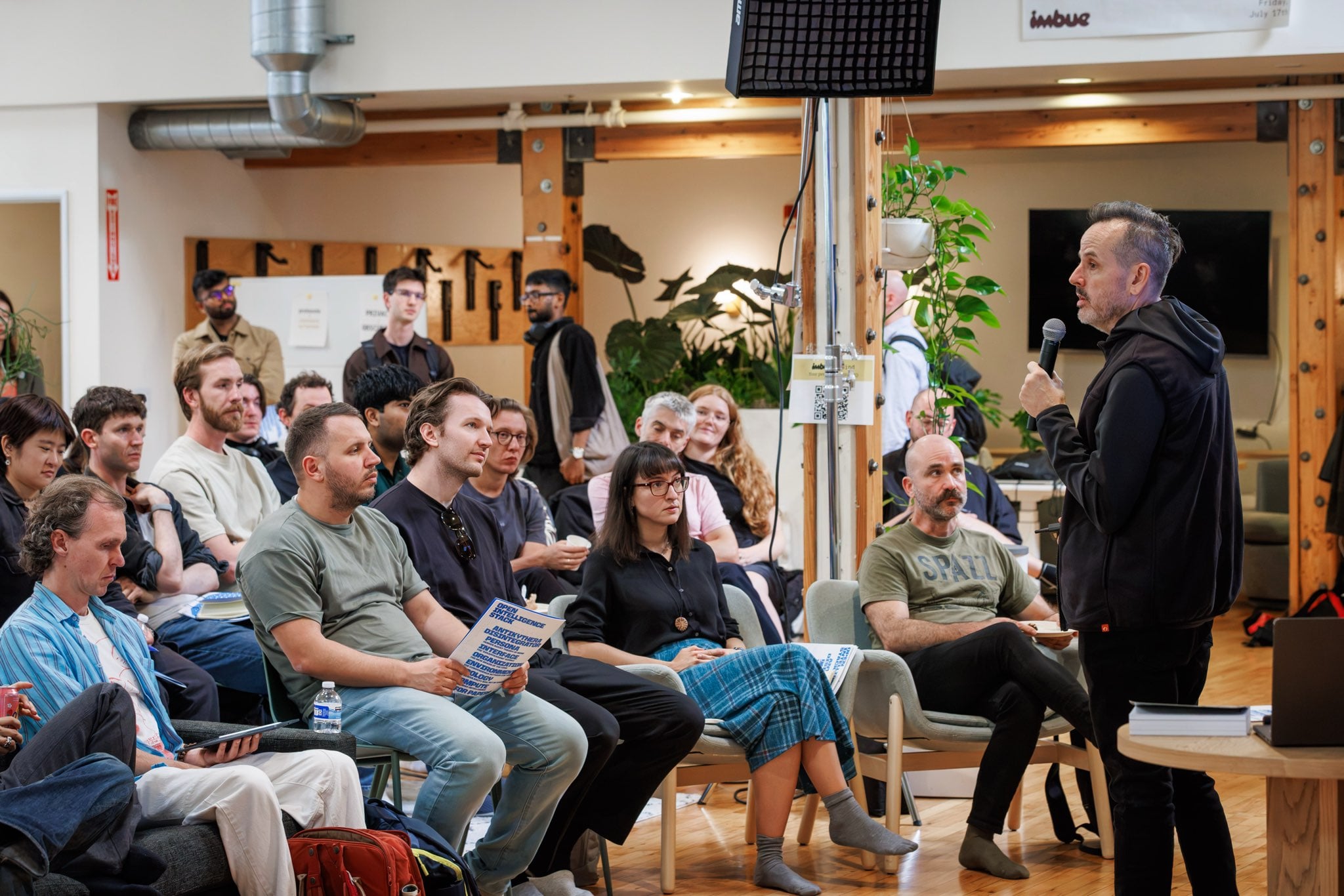 A crowded room at the Antikythera x Disintegrator salon, listening to someone speak into a microphone; a handout titled Open Intelligence Stack, listing Persona and Interface among its sessions, is visible in the audience.